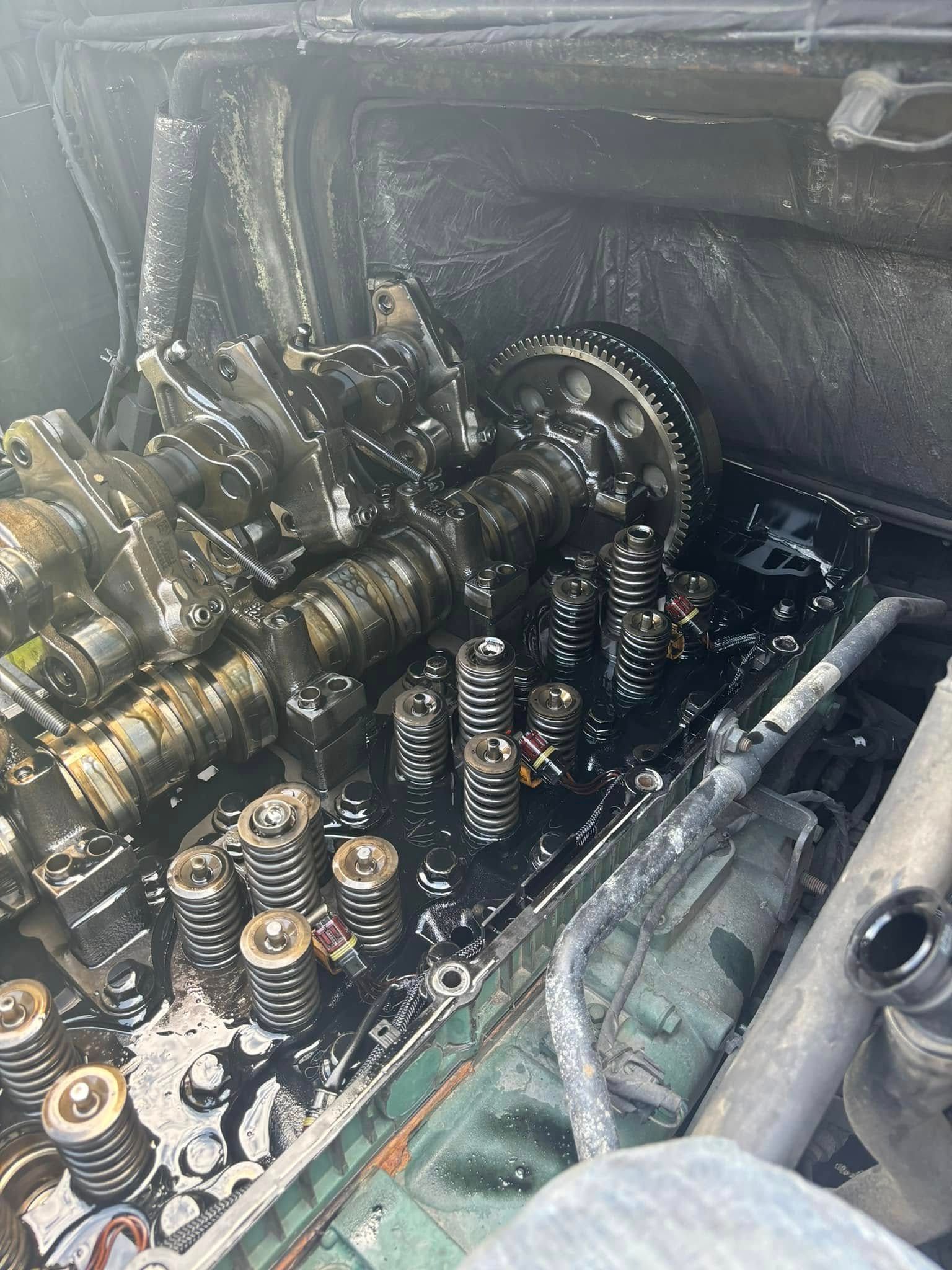 An open, oil-coated car engine cylinder head showing the camshaft, valve springs, and dark, sludge-covered components.