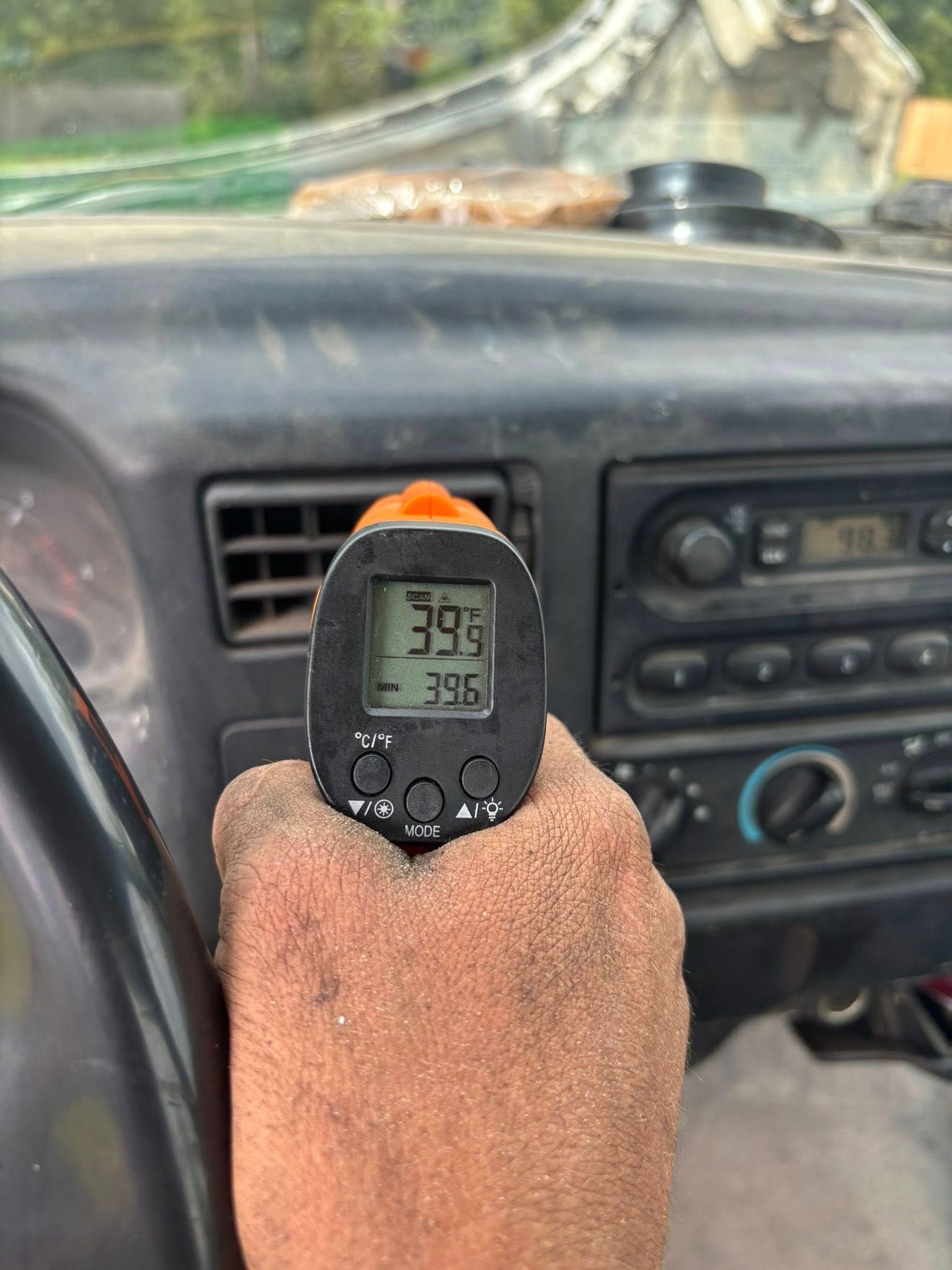 A person’s hand holds an infrared thermometer displaying 39 degrees in front of a vehicle's dashboard air vent.
