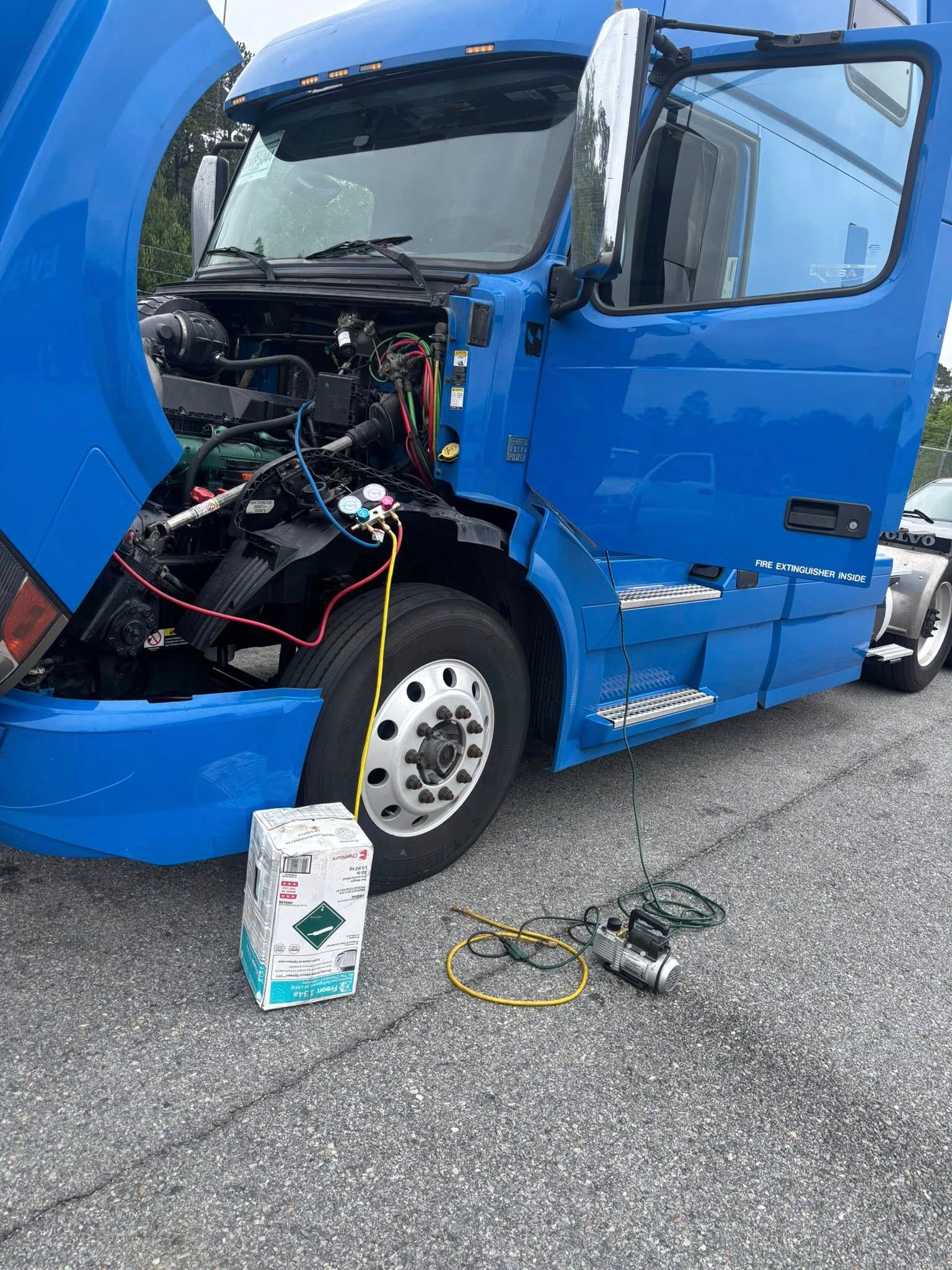 A blue semi-truck with an open hood is being serviced with a refrigerant tank and tools on the ground.