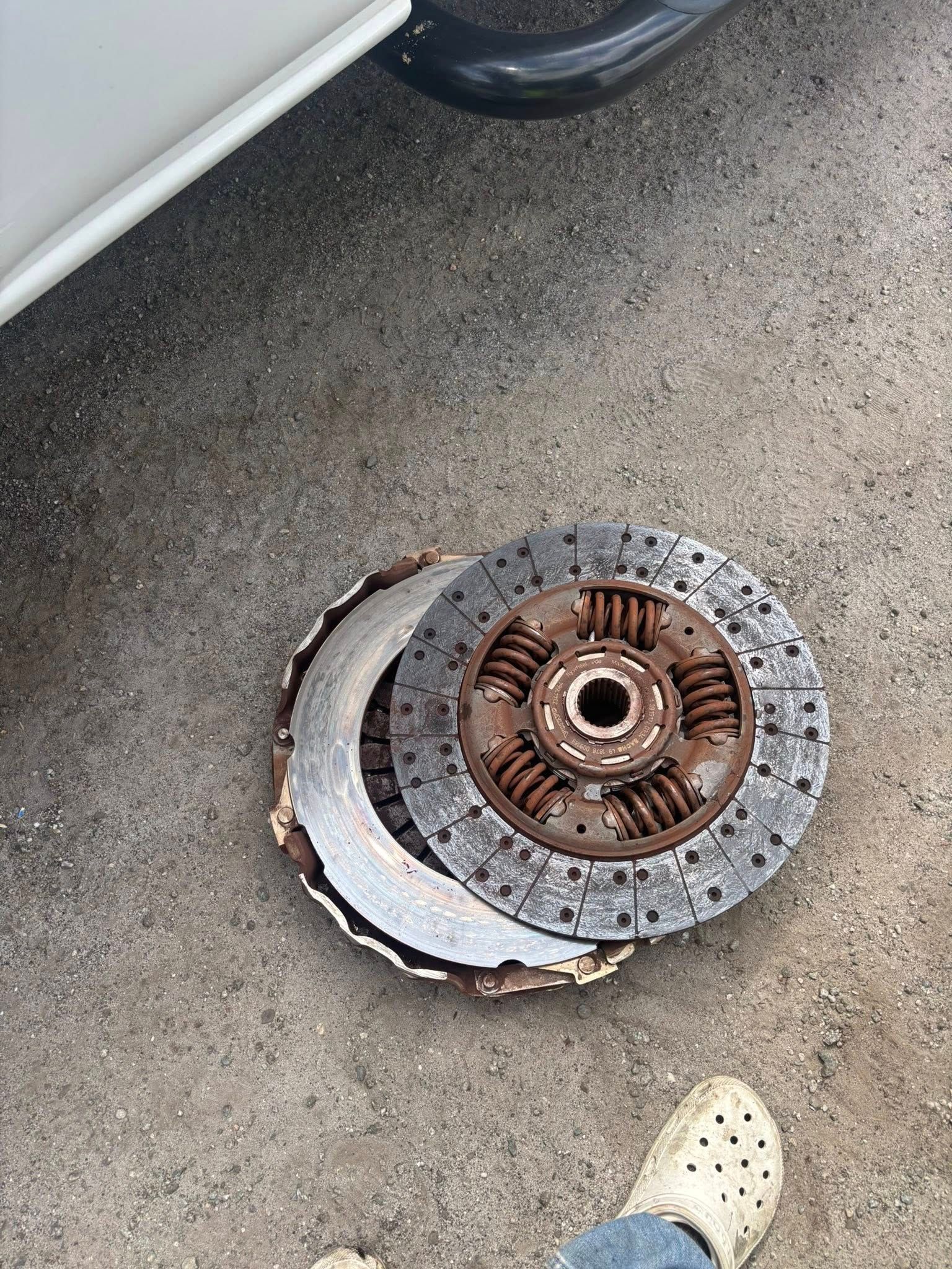 A worn and damaged automotive clutch disc lying on the pavement next to a vehicle.