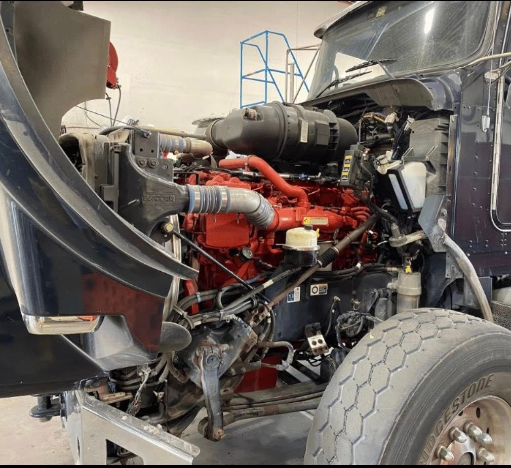 The open hood of a dark blue semi-truck reveals a large, red engine with various hoses and components in a shop setting.