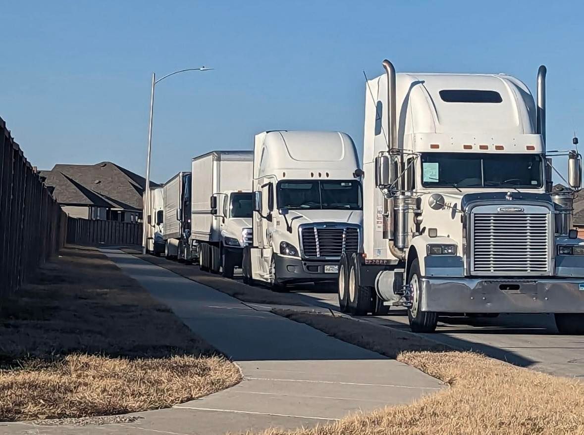 A line of white semi-trucks parked single-file along a suburban street curb next to a residential fence.