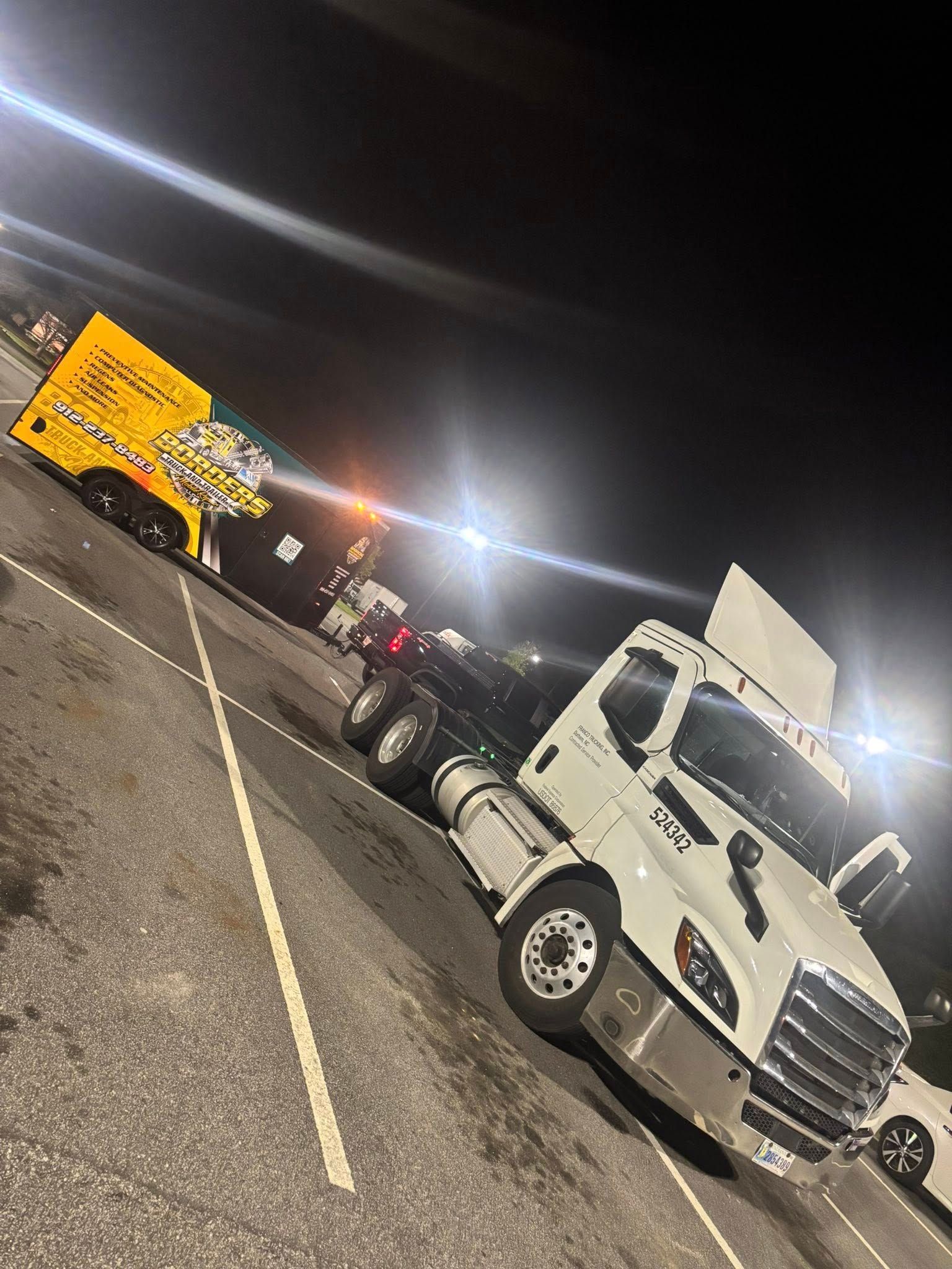 A white semi-truck parked at night, attached to a yellow trailer with a logo, in a parking lot.