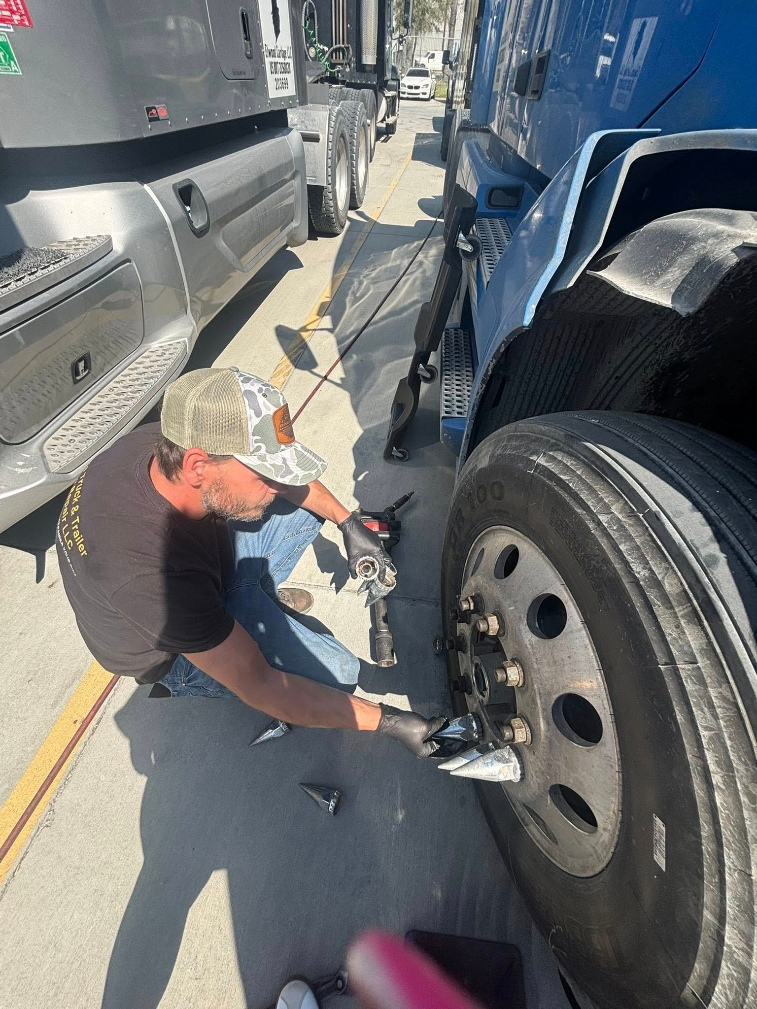 A person wearing a cap and gloves kneeling to work on the lug nuts of a semi-truck tire on an asphalt surface.