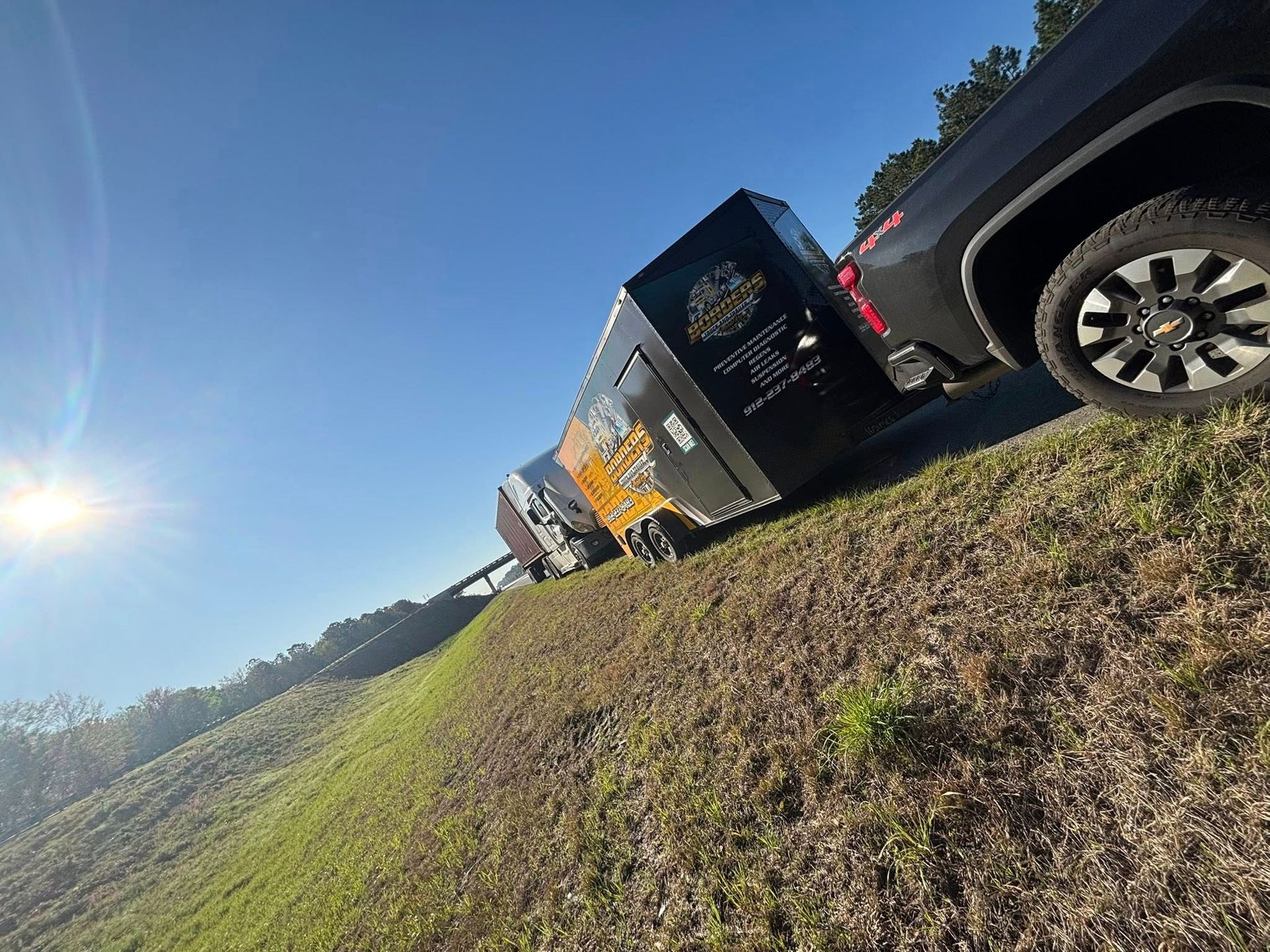 A black pickup truck towing an enclosed trailer parked on a grassy embankment under a bright, sunny sky.