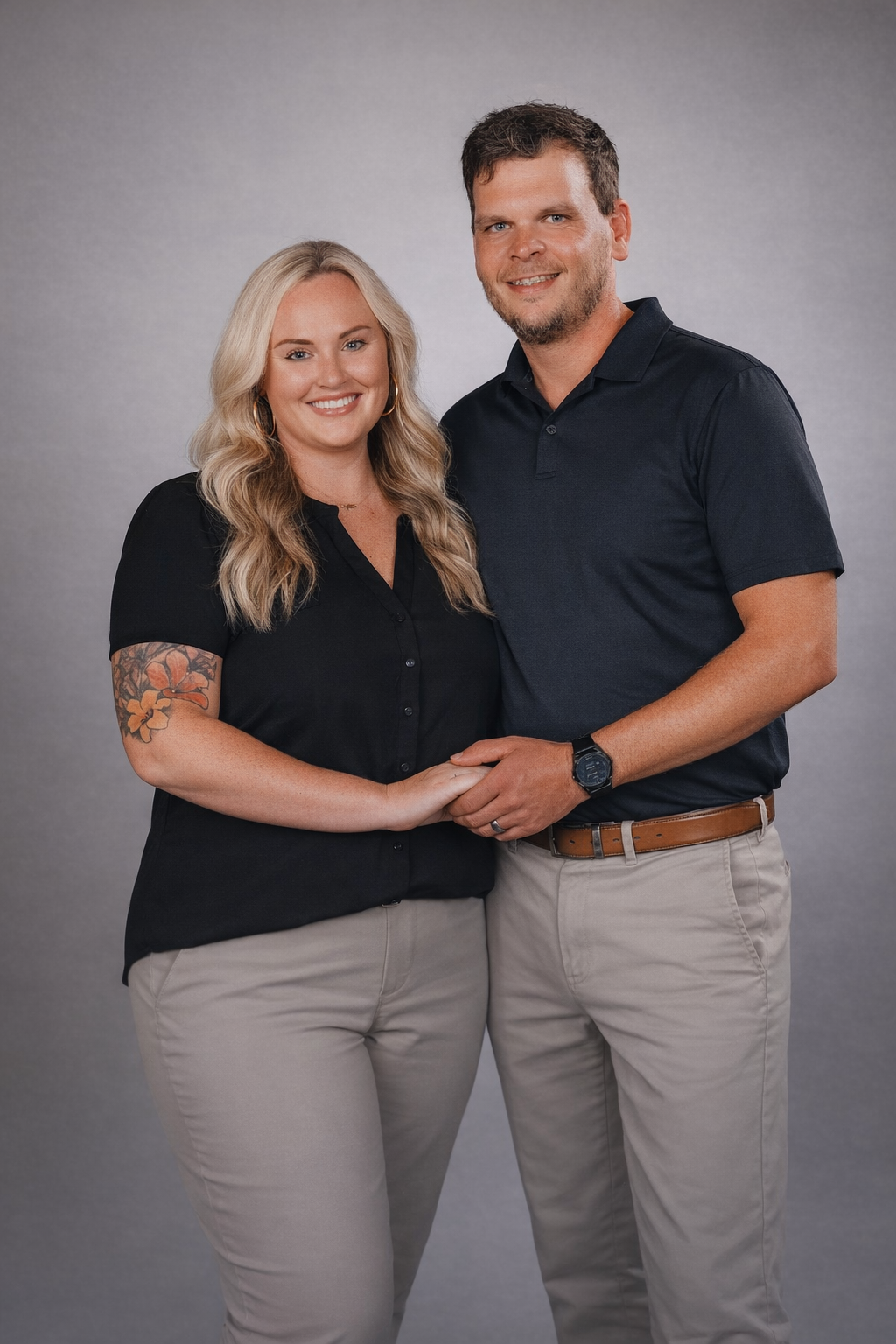 A man and a woman in matching black shirts and khaki pants stand together smiling against a grey studio backdrop.