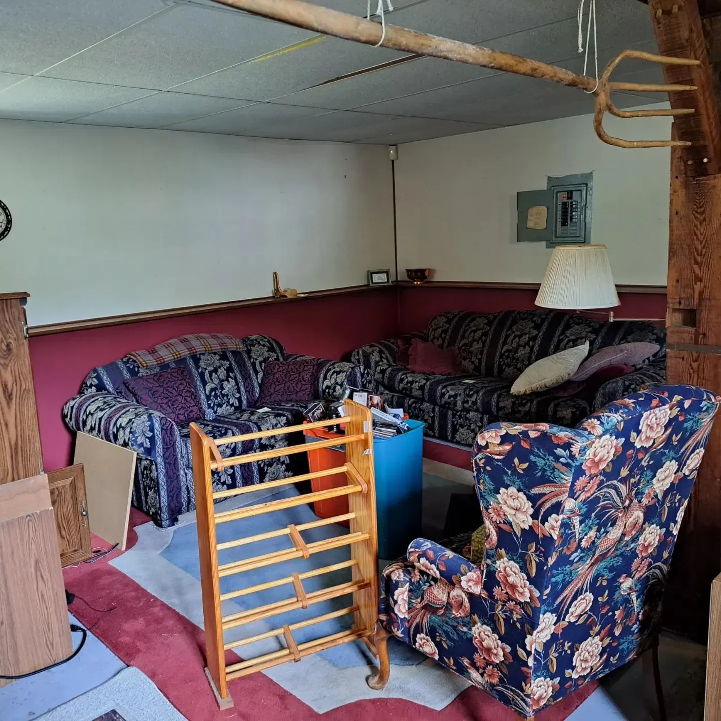 A cluttered living room with two sofas, an armchair, and a wooden rack. Dark red and white walls.