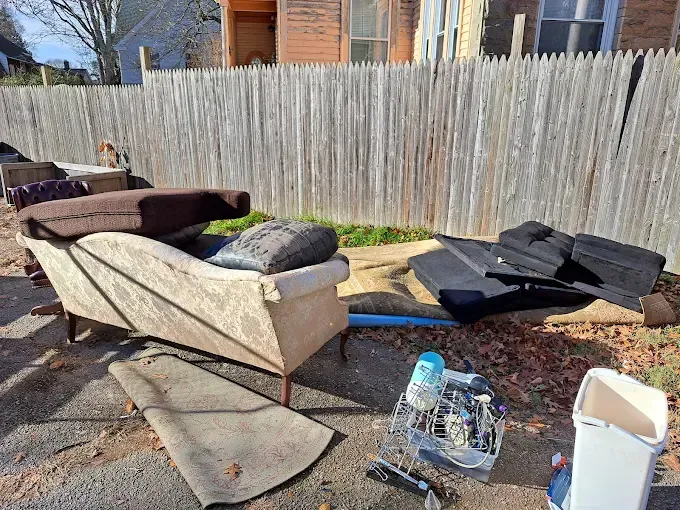 Pile of discarded furniture and debris on the side of a road, next to a wooden fence.