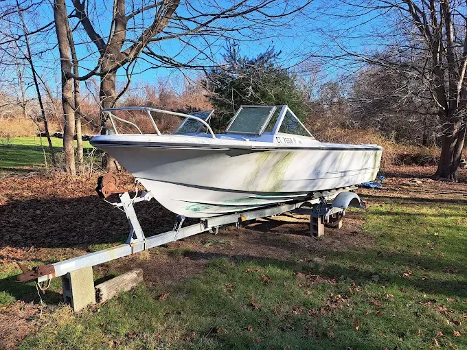 Boat on a trailer in a grassy yard, surrounded by bare trees under a blue sky.
