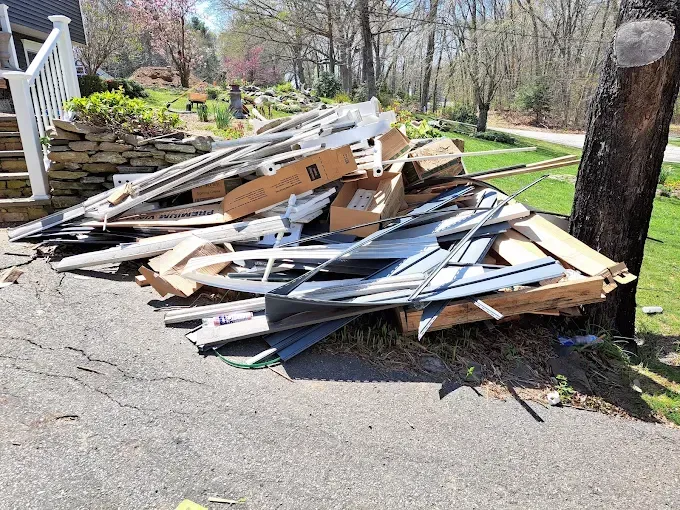 Pile of construction debris and cardboard boxes on a driveway next to a tree and house.