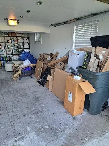 Cluttered garage interior. Boxes, trash, shelves with items, and a fire pit are visible against a concrete floor.