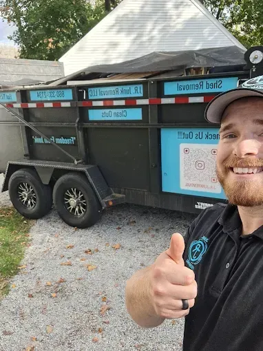 Man smiling, giving thumbs-up, next to a black dumpster trailer with teal lettering and logo.