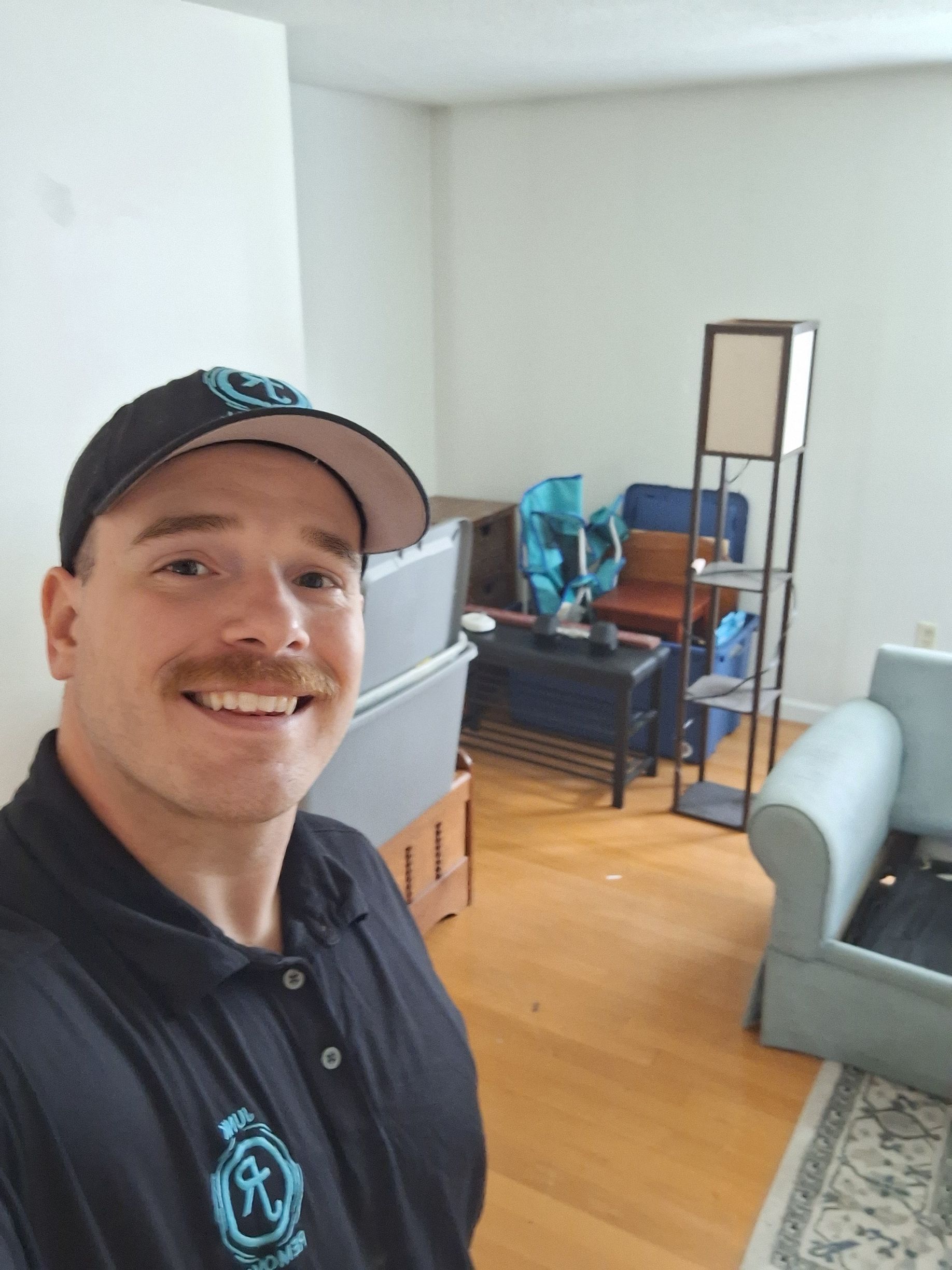 Smiling man in a black cap and shirt taking a selfie in a bright living room with a gray sofa and mirror cabinet.
