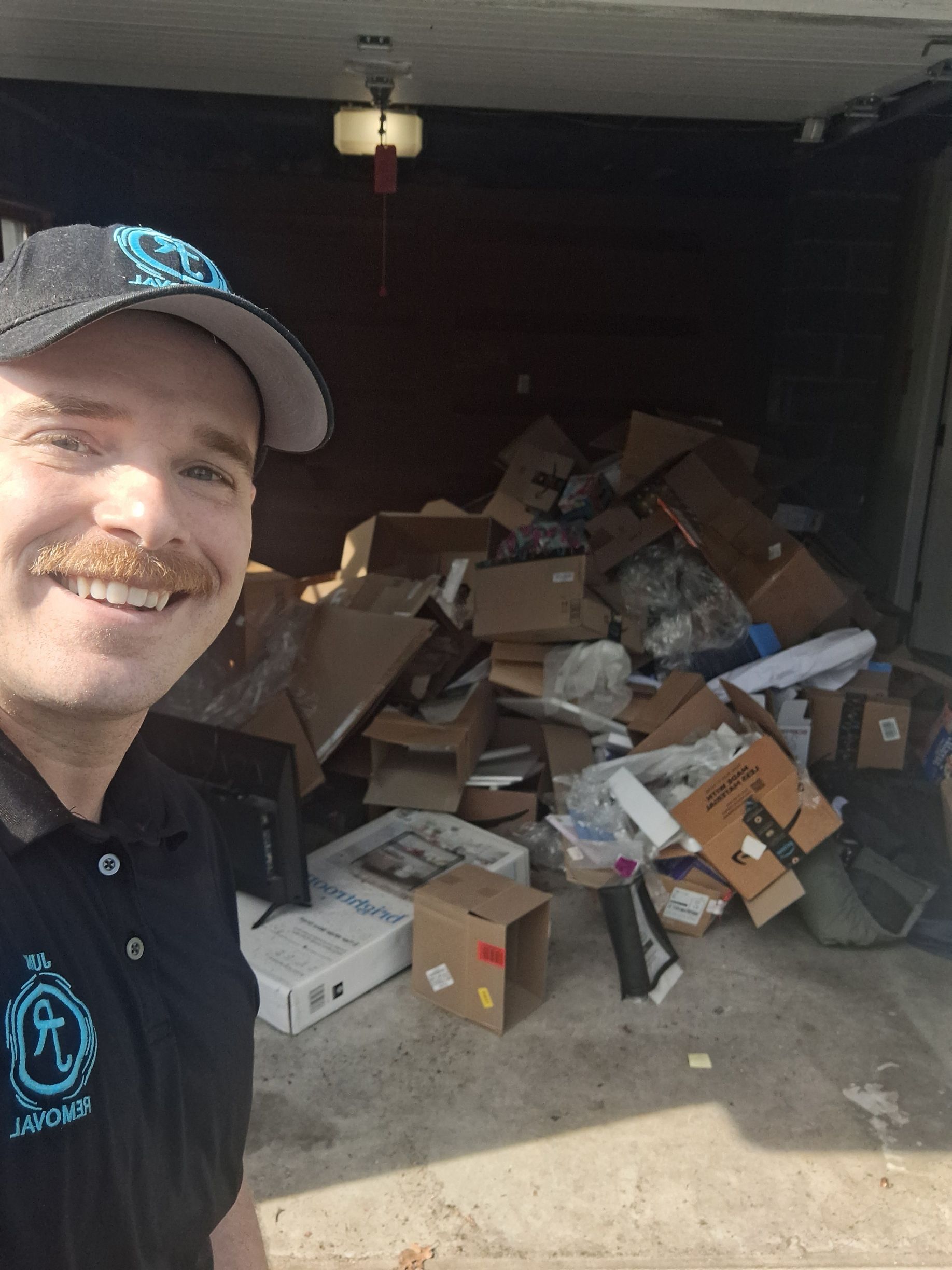 Smiling man in black polo and cap selfie beside a garage full of cardboard and trash bags