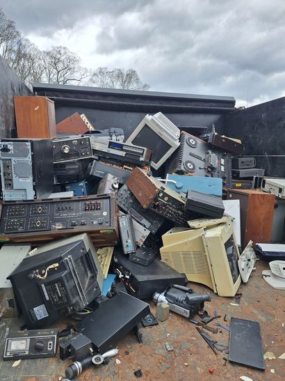 Pile of old electronics including CRT monitors, computers, and a refrigerator on a wooden pallet in a yard.