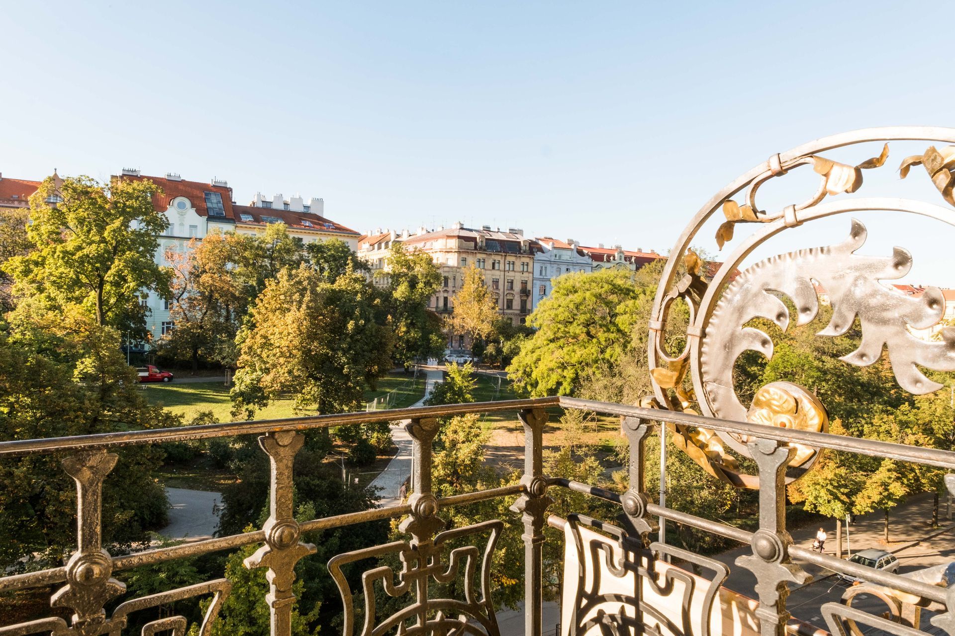 View from ornate balcony overlooking a park and buildings on a sunny day.