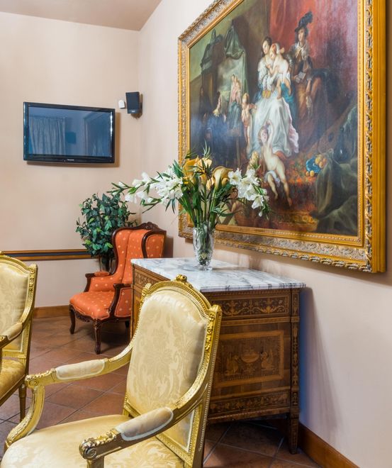 Ornate room with chairs, antique cabinet, large painting, flowers, and television on the wall.