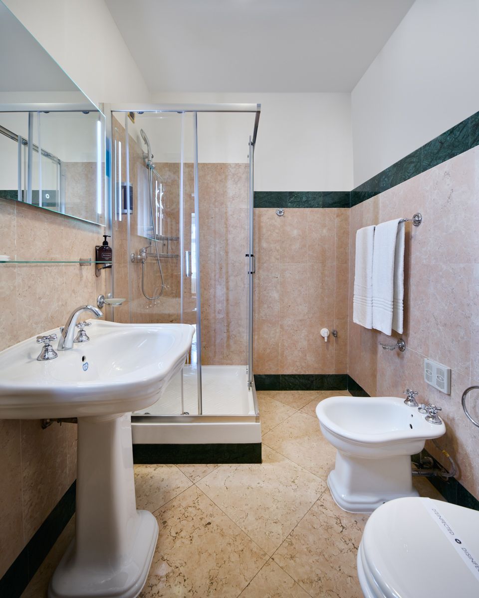 Bagno con lavabo su piedistallo, doccia in vetro e bidet. Dettagli beige e verdi.