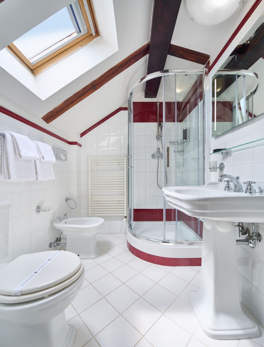Bagno con piastrelle bianche, lucernario e doccia in vetro. Dettagli rossi, WC, bidet e lavabo a colonna.