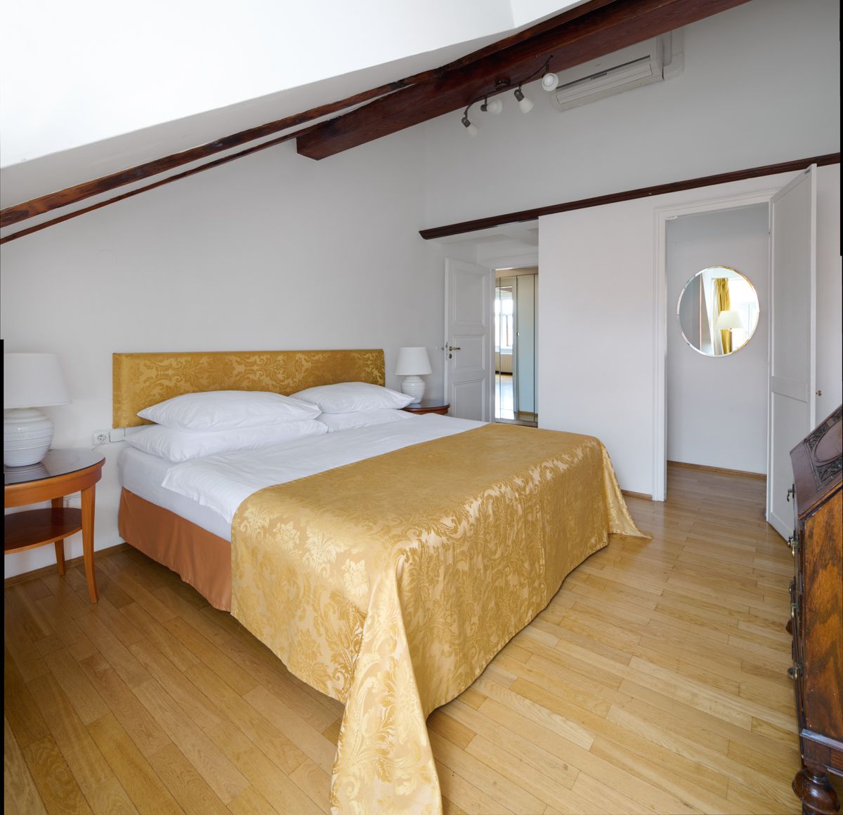 Camera da letto con pavimenti in legno, letto con copriletto dorato, pareti bianche e un piccolo tavolino.