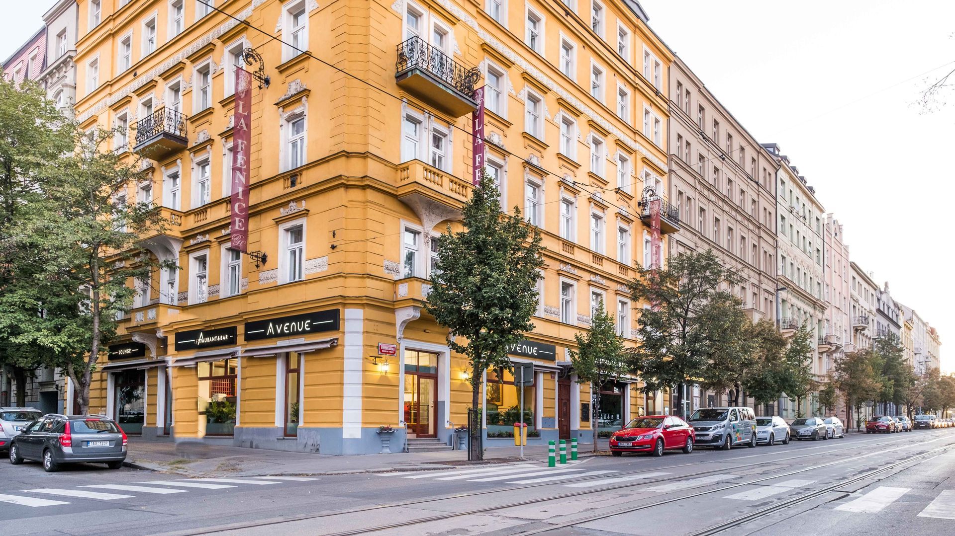 Edificio giallo con vetrine su una strada cittadina, auto parcheggiate, alberi lungo il marciapiede, binari del tram.