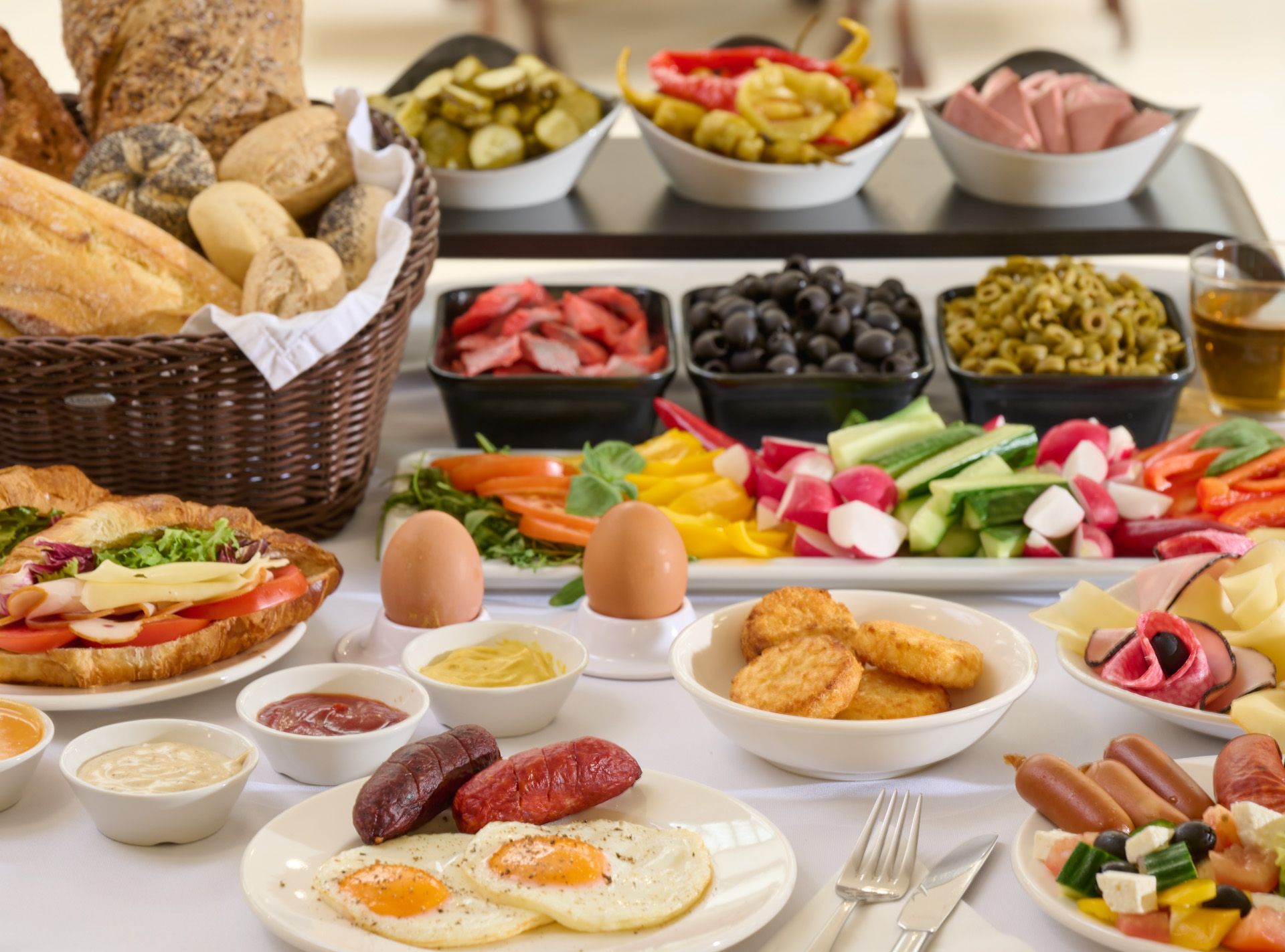 Colazione a buffet con vari tipi di pane, carne, verdure, uova e contorni serviti su una tovaglia bianca.