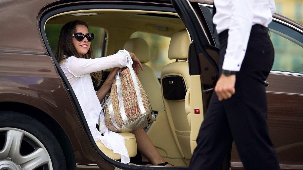 Woman in sunglasses seated in a brown luxury car, holding a bag, and smiling as the door is opened.