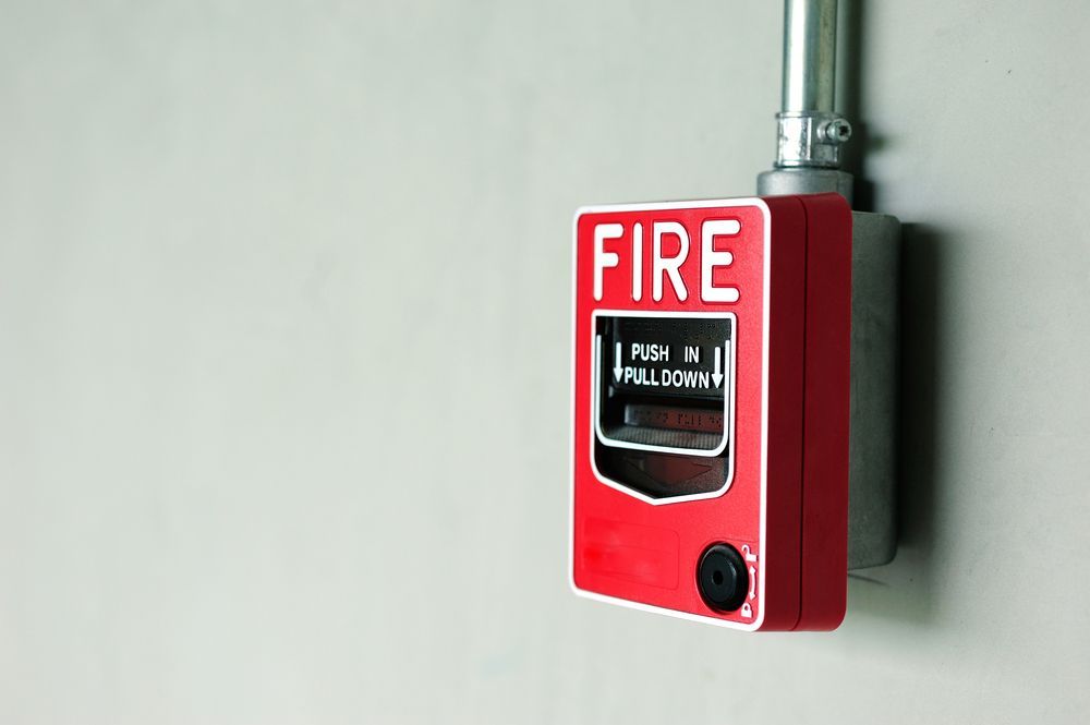 A red, rectangular fire alarm pull station mounted on a plain light-gray wall with a metal conduit pipe above it.