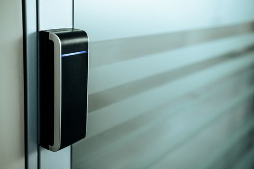 A black and silver electronic access control reader mounted on a metal door frame next to a frosted glass door.
