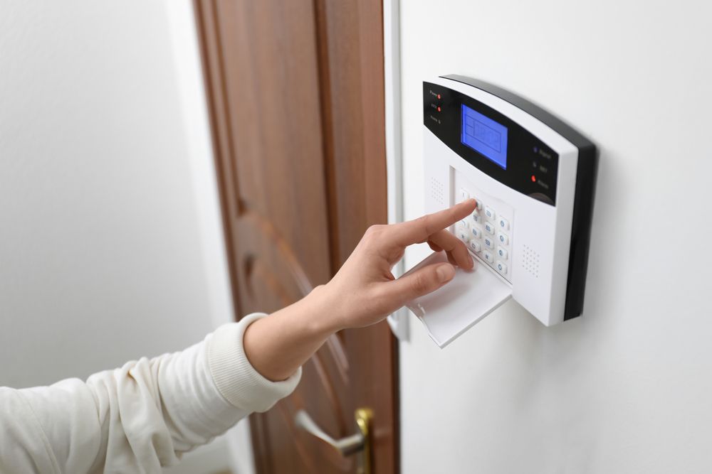 A person's hand enters a code on a white home security keypad mounted on a wall next to a wooden door.