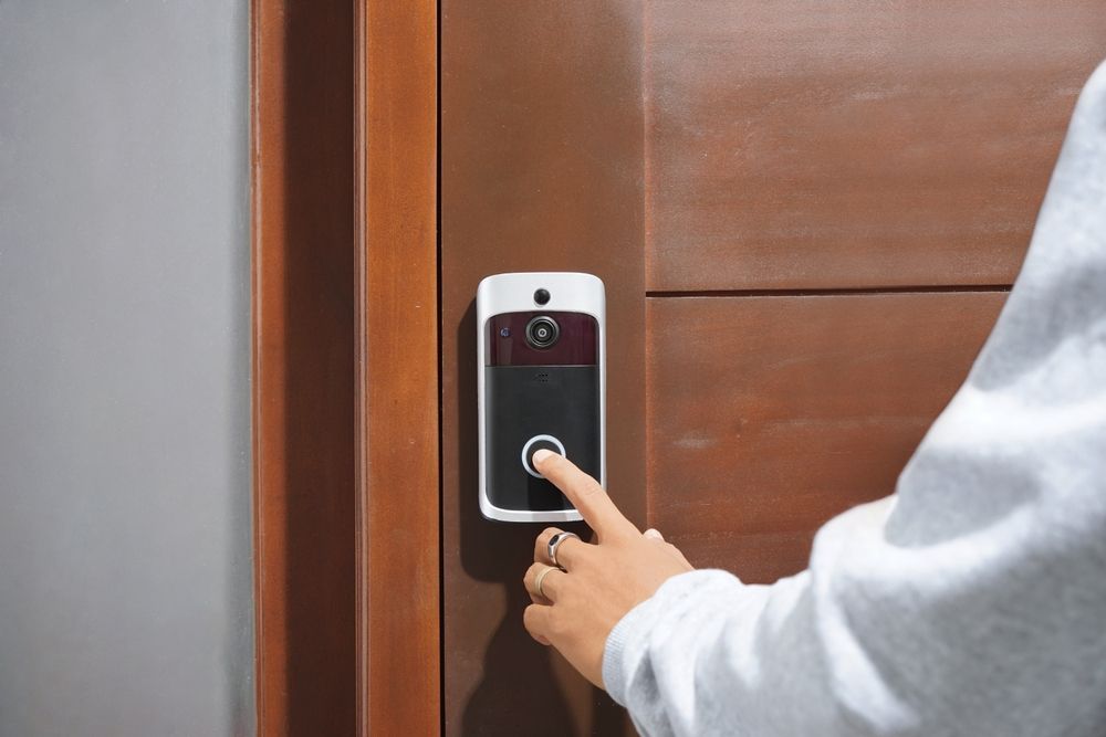 A hand in a grey sleeve presses the button on a smart video doorbell mounted to a wooden door.