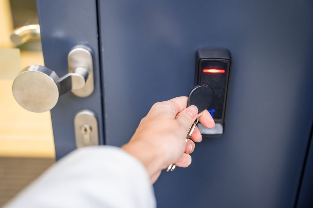 A hand holds a key fob against a wall-mounted electronic card reader next to a door handle on a blue door.