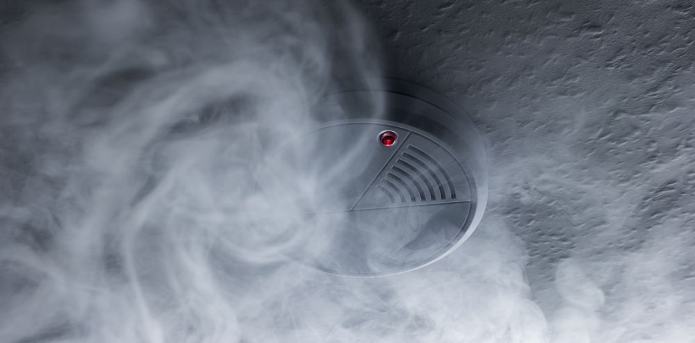 A smoke detector mounted on a ceiling is surrounded by swirling white smoke.