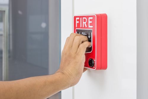 A hand pulls down on a red fire alarm station mounted on a white wall.