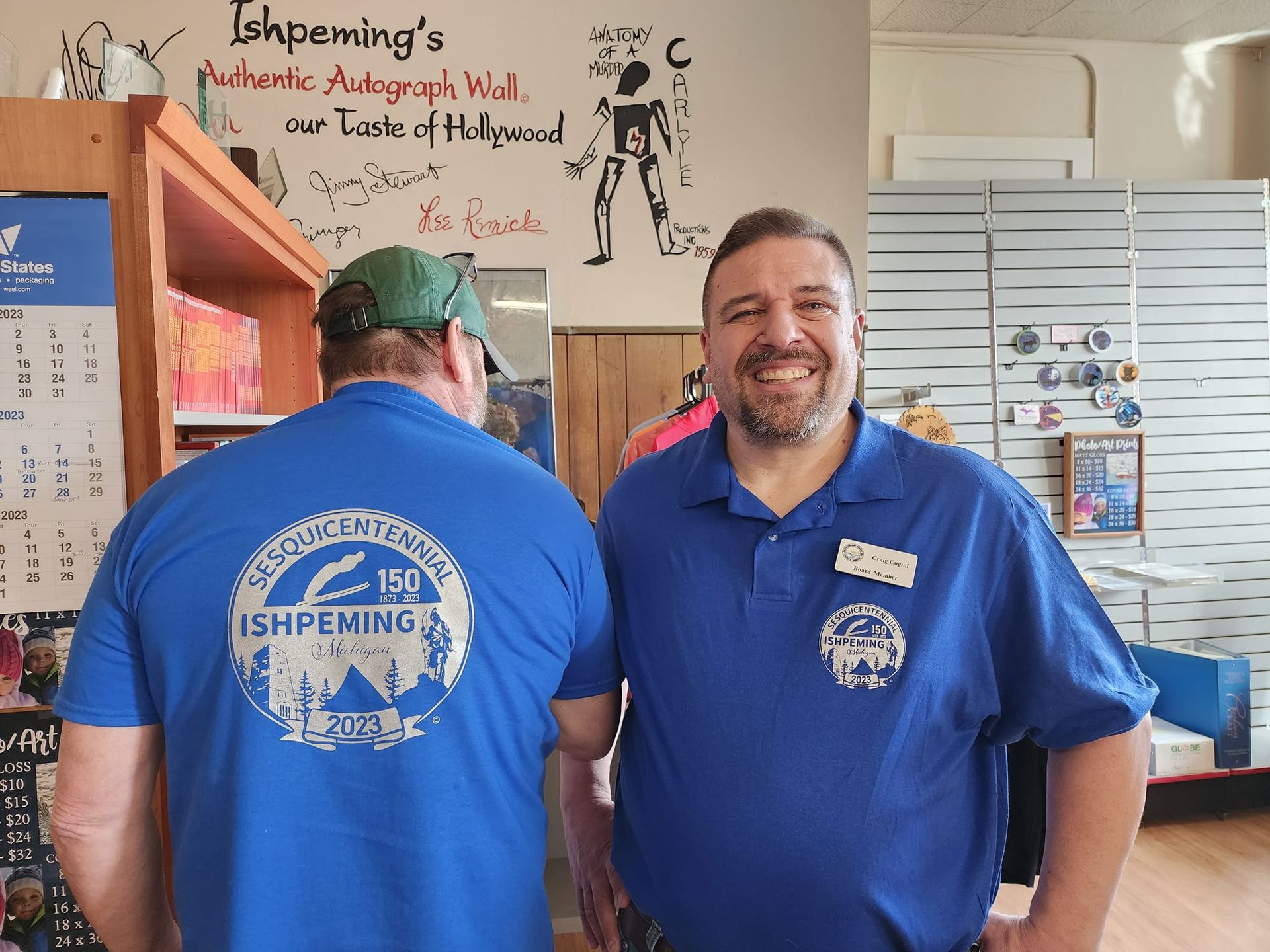 Two men wearing blue shirts with a logo, smiling in a store with an autograph wall.