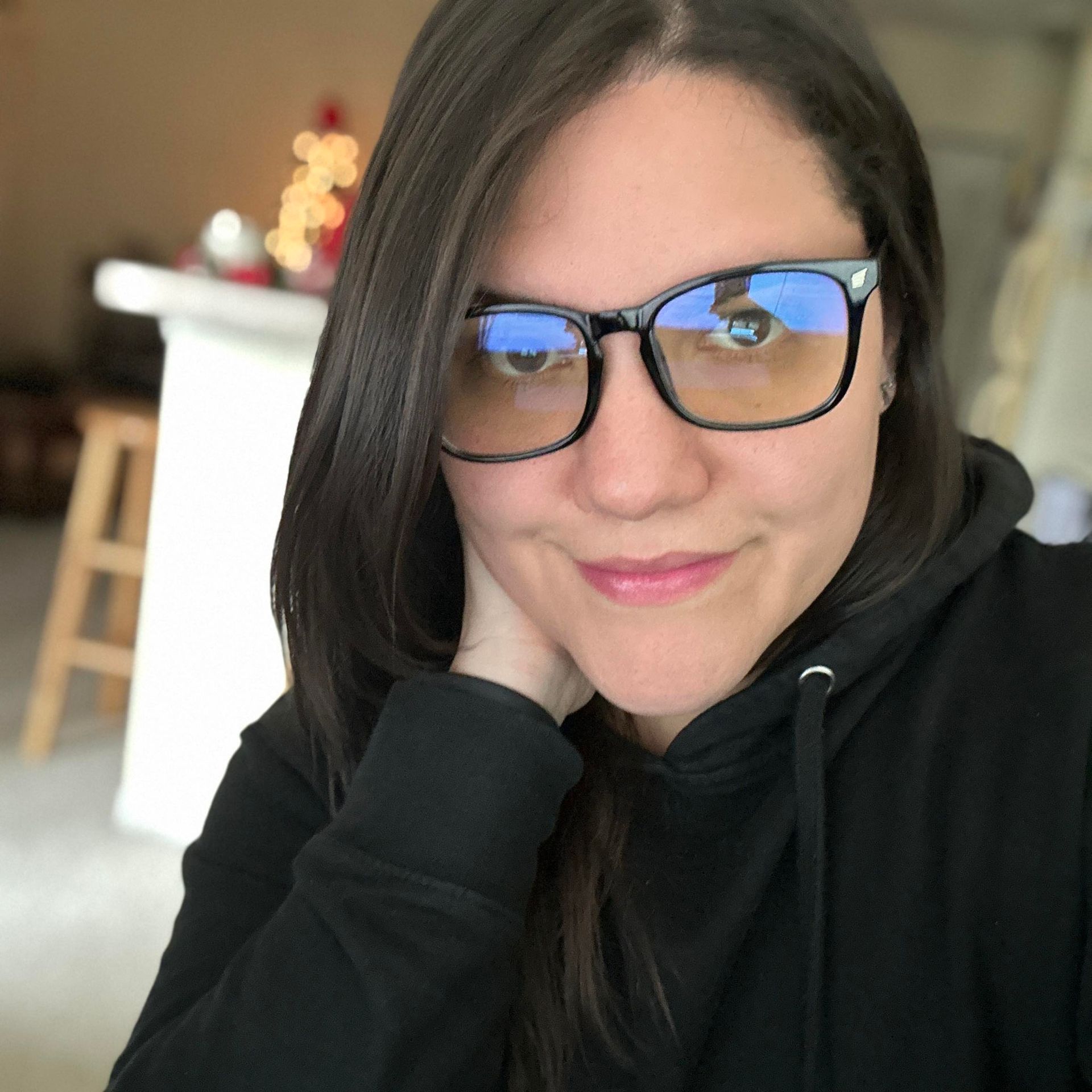 Woman with dark hair and glasses in a black hoodie, resting chin on hand; soft smile.