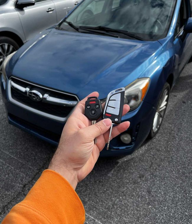 Hand holding a Subaru car key and fob in front of a blue Subaru in a parking lot