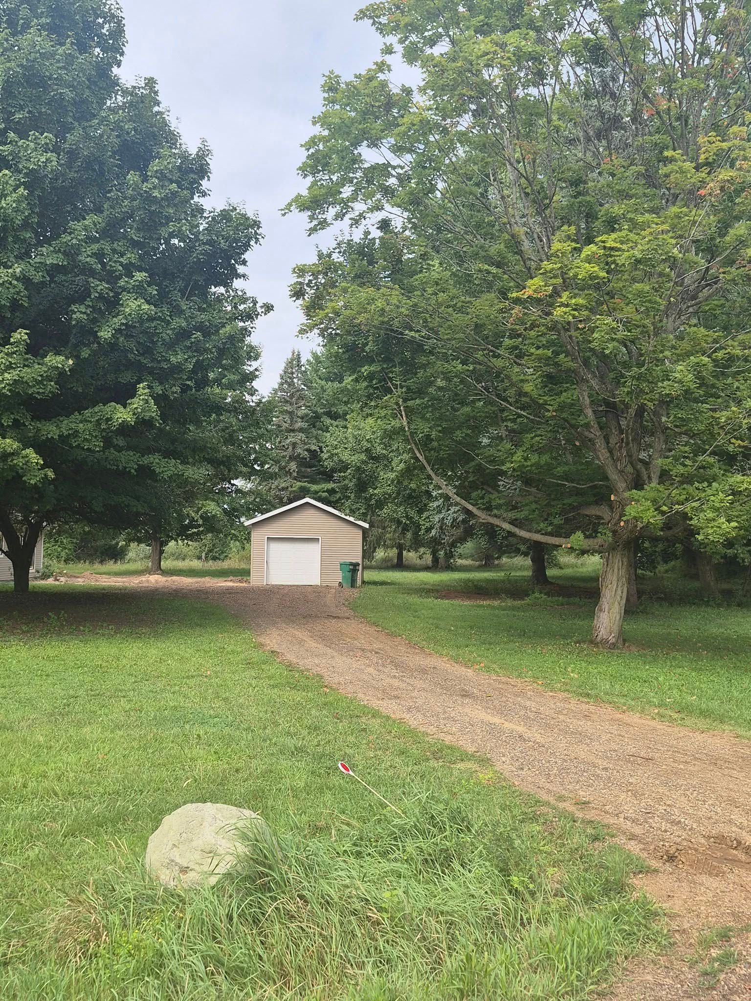 Gravel driveway leads to a small, beige garage, flanked by trees. A rock sits in the grassy foreground.