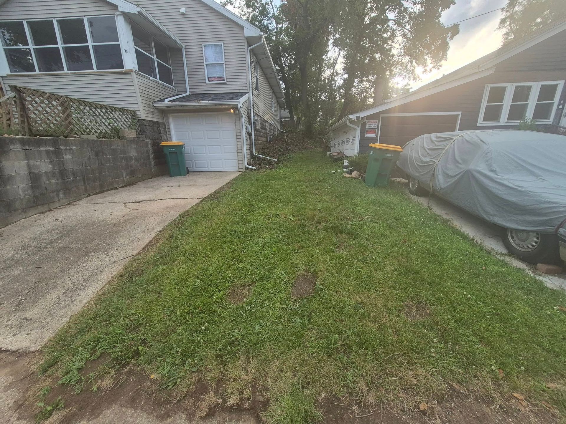 Lawn between houses, overgrown with weeds. Driveway and parked car visible. Green trash bins on each side.