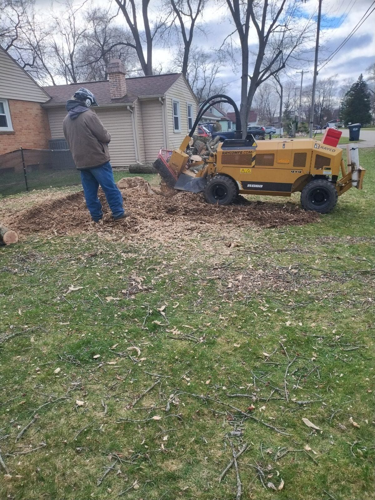 Man using a yellow stump grinder in a yard, making wood chips. Houses and trees are in the background.
