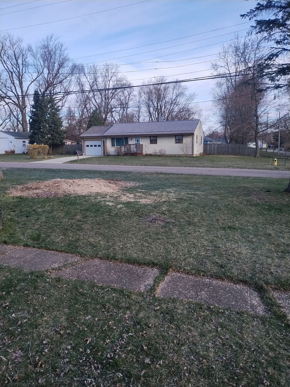 A small, beige house with a garage sits behind a lawn. Bare trees and a blue sky in background.