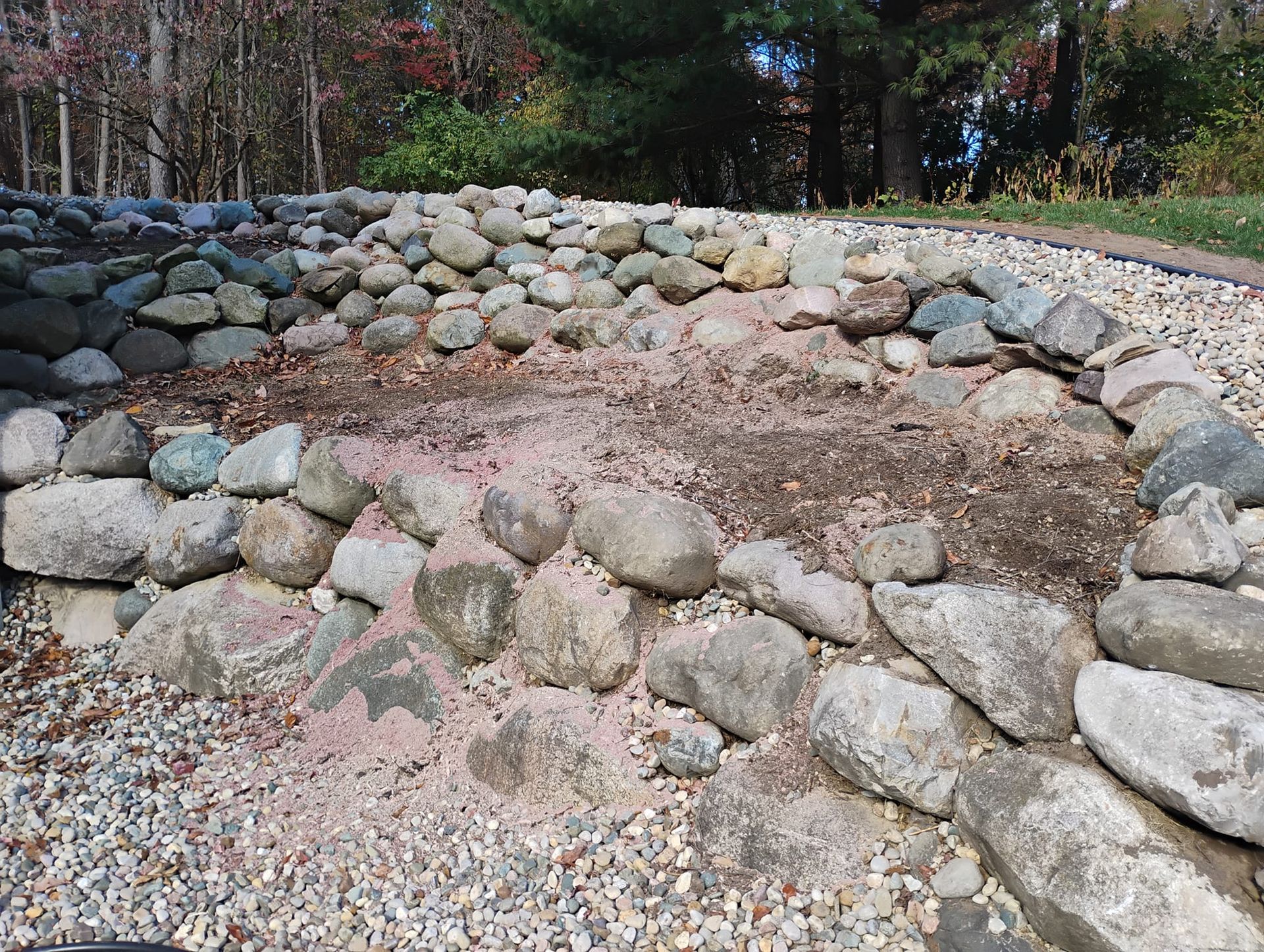 Stone-lined garden bed filled with dirt and gravel, surrounded by rocks; outdoors, in sunlight.
