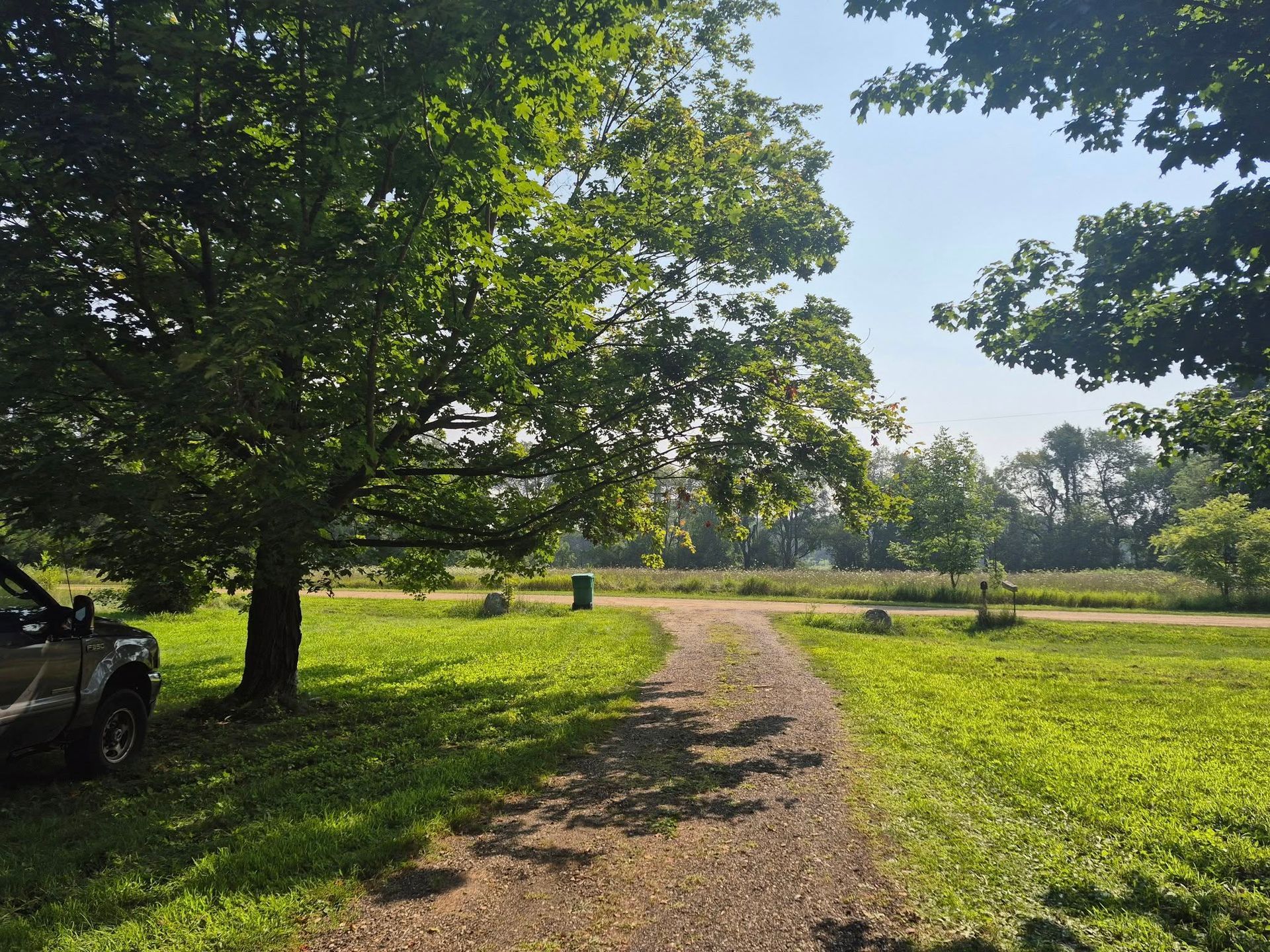 Gravel path through a grassy park, shaded by large trees. A vehicle is parked near the tree on the left.