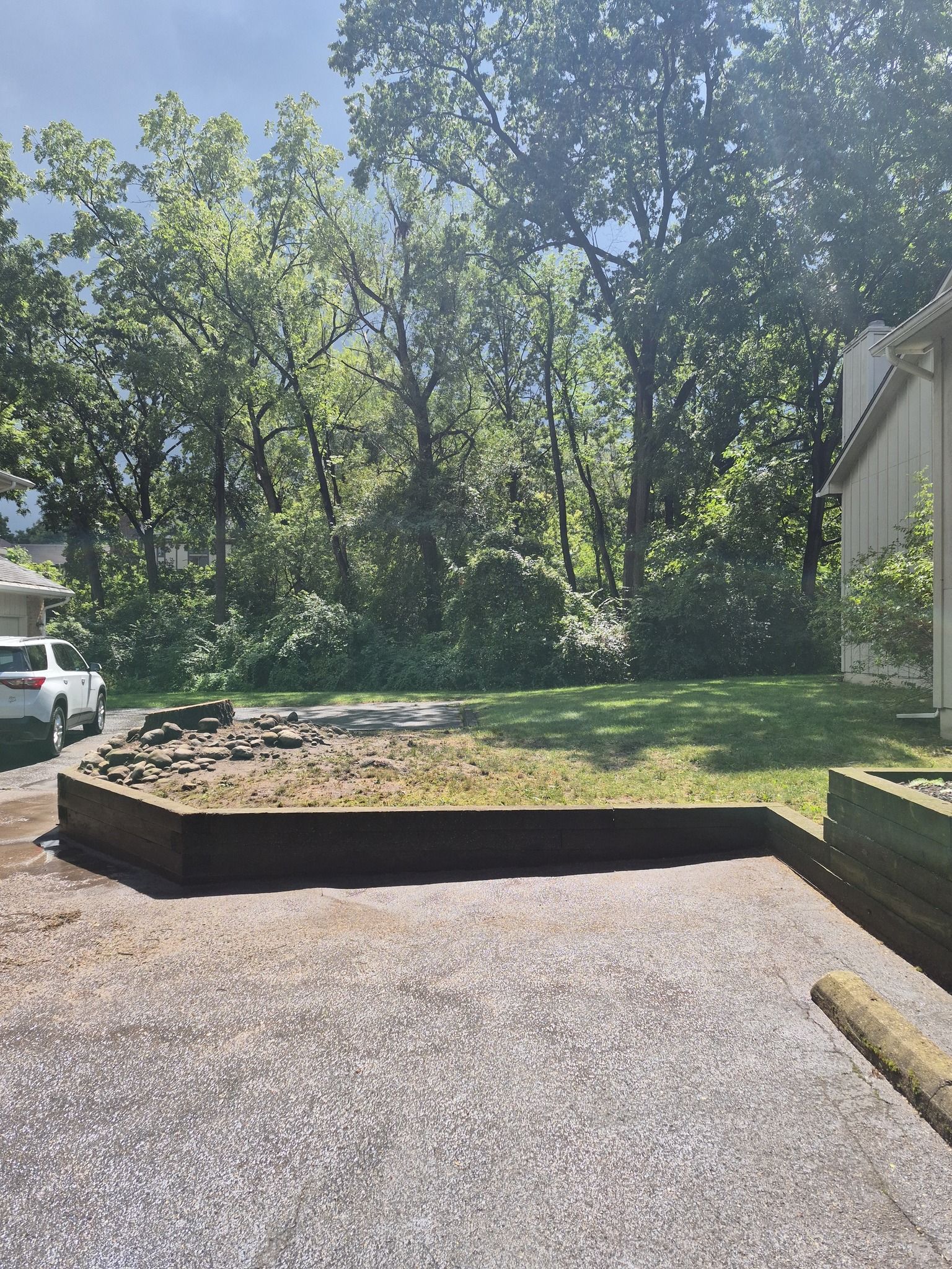 Asphalt area in front of a raised planter with a car to the left and trees in the background.