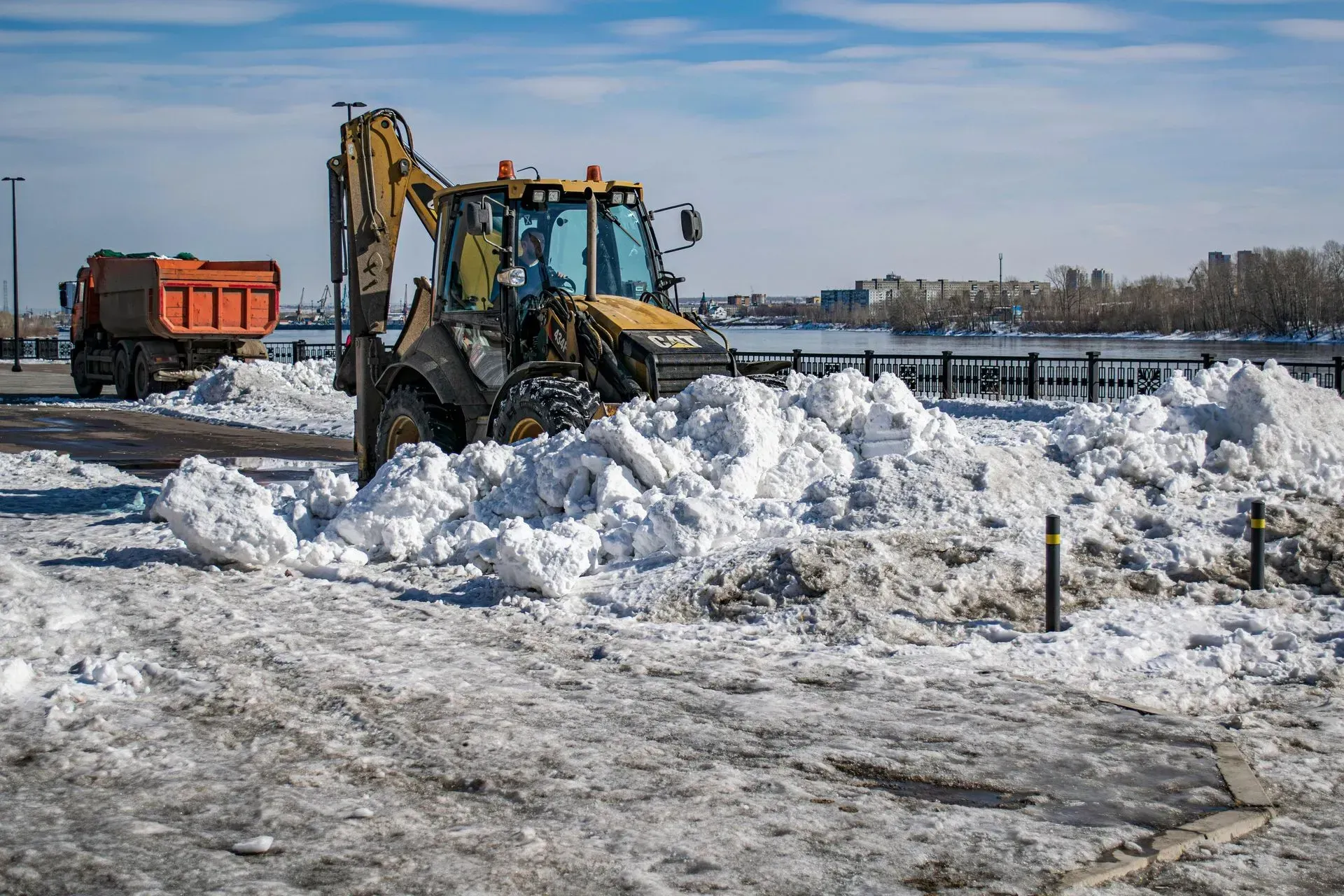 Yellow backhoe clearing snow, orange dump truck in background, sunny day, cityscape in distance.