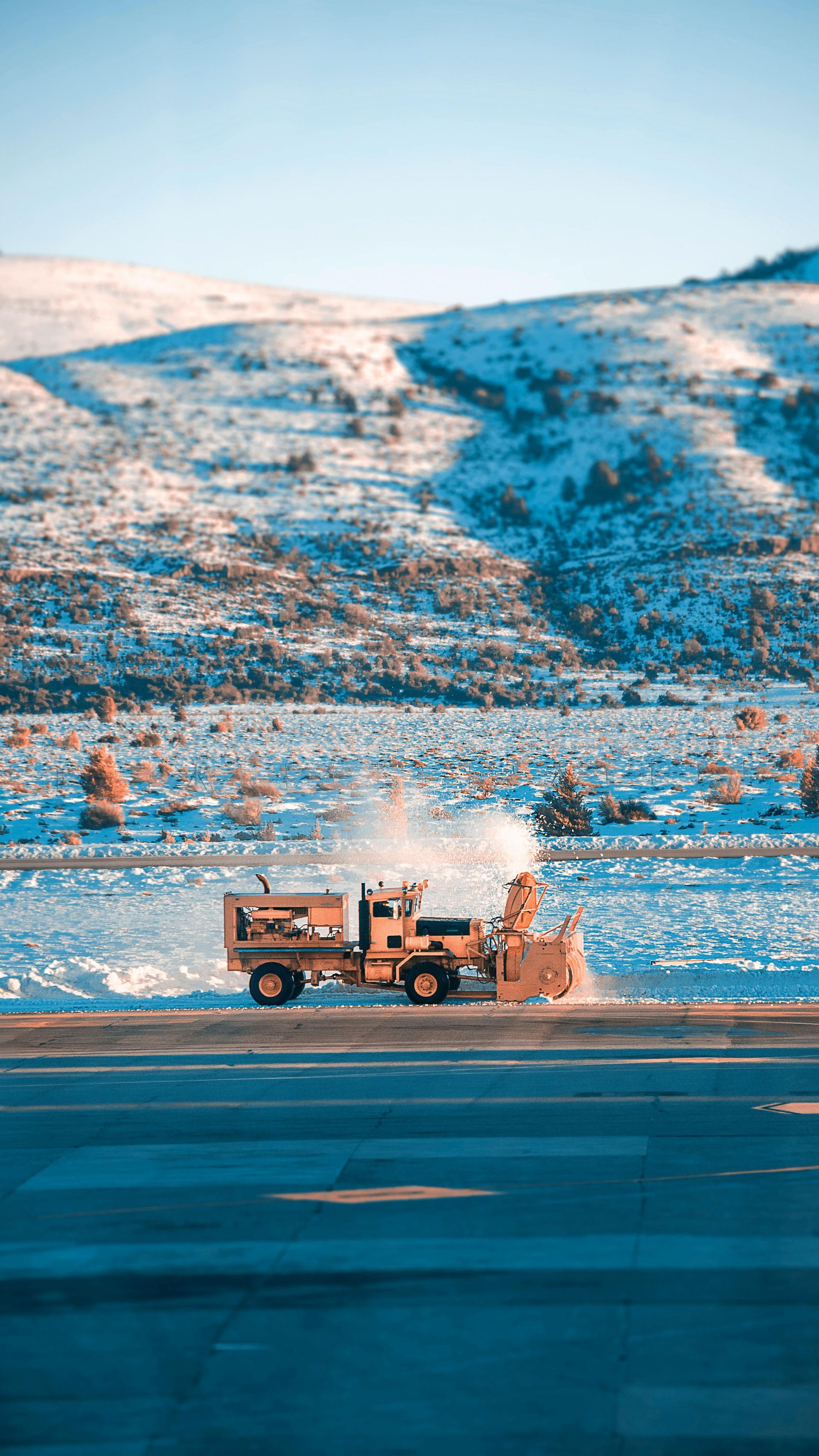 Yellow snowblower clearing a road in front of snow-covered mountains, blue sky.