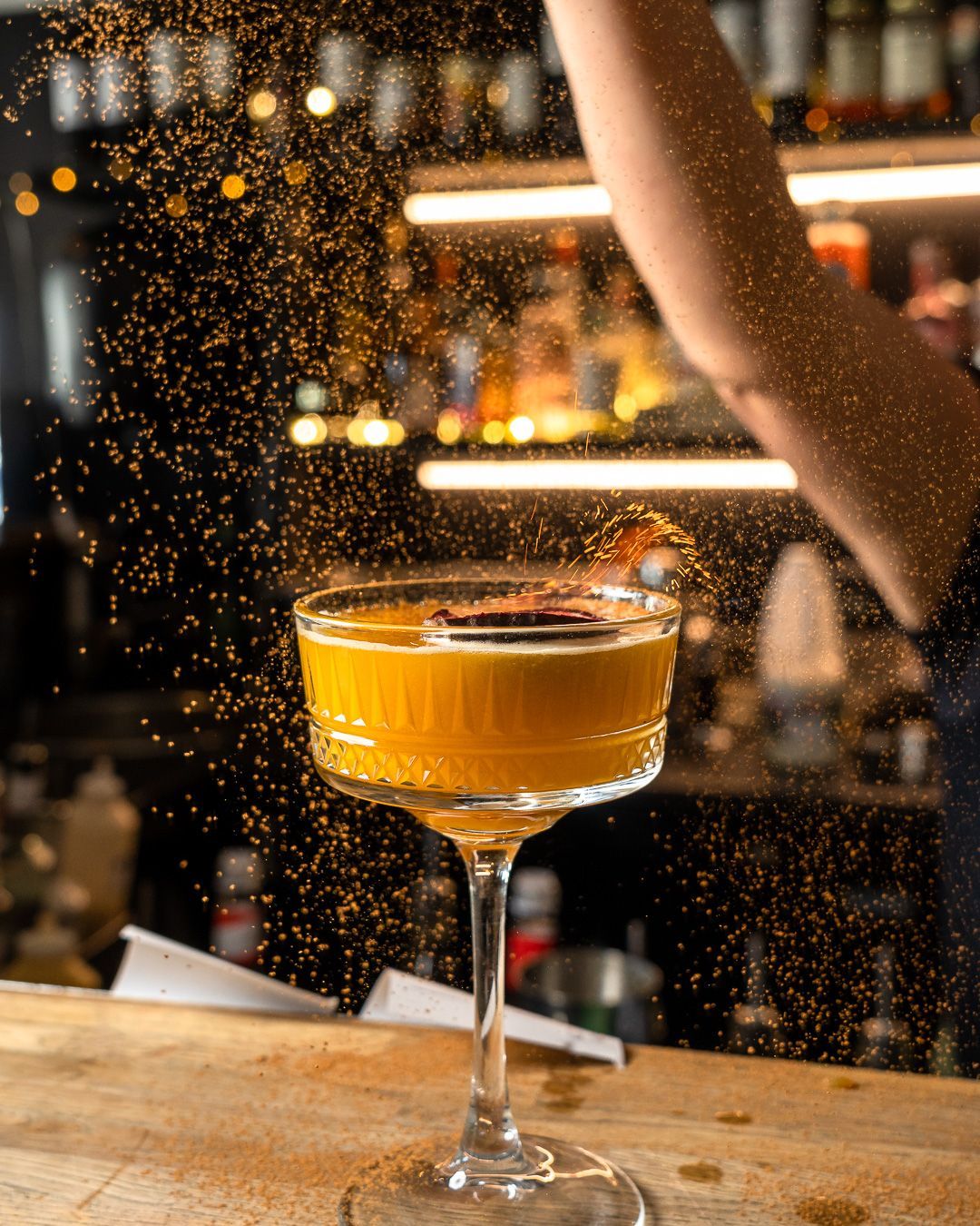 Cocktail being garnished with a spice; bartender's arm visible. Orange drink in a stemmed glass on a bar.