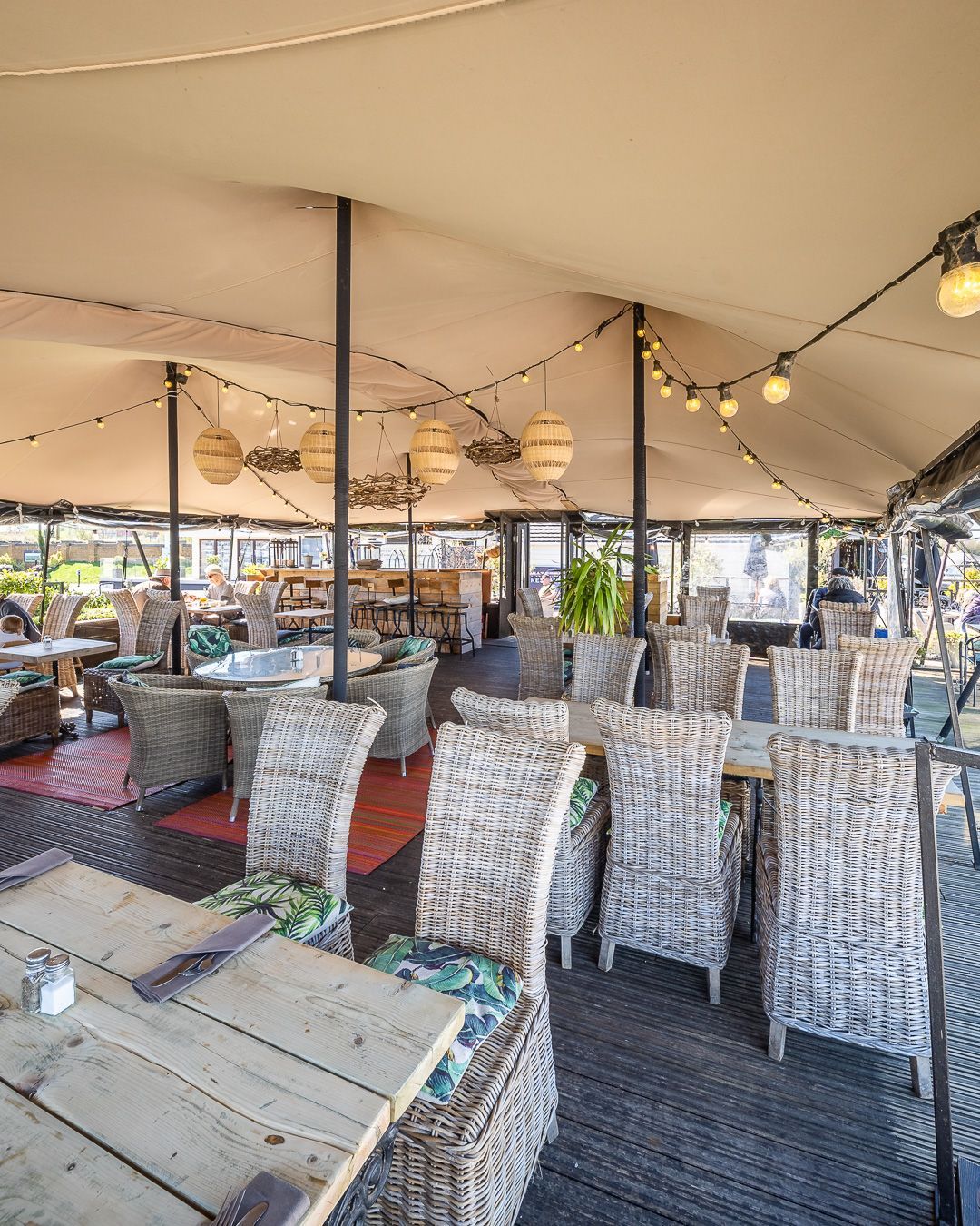 Outdoor restaurant seating area with woven chairs, tables, and string lights under a tent.