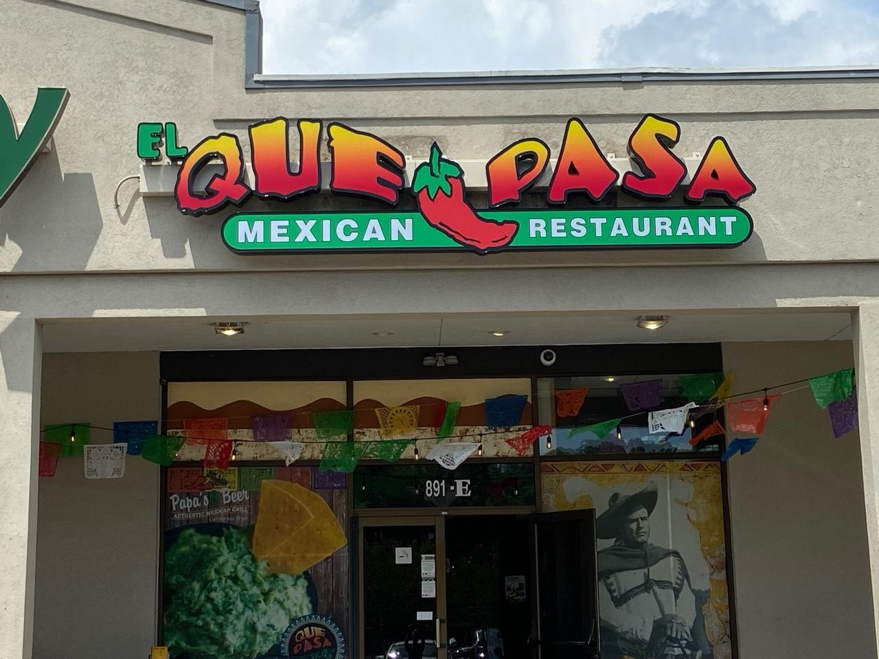 El Que Pasa Mexican Restaurant sign with colorful letters and a chili pepper over a doorway with flags and banners.