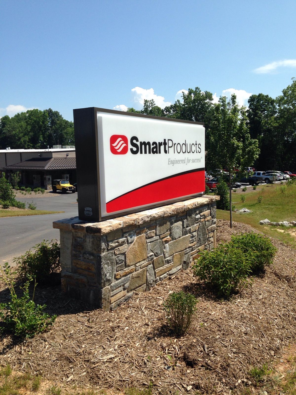 Sign for Smart Products, with logo over a stone base, in front of a building and trees under a blue sky.